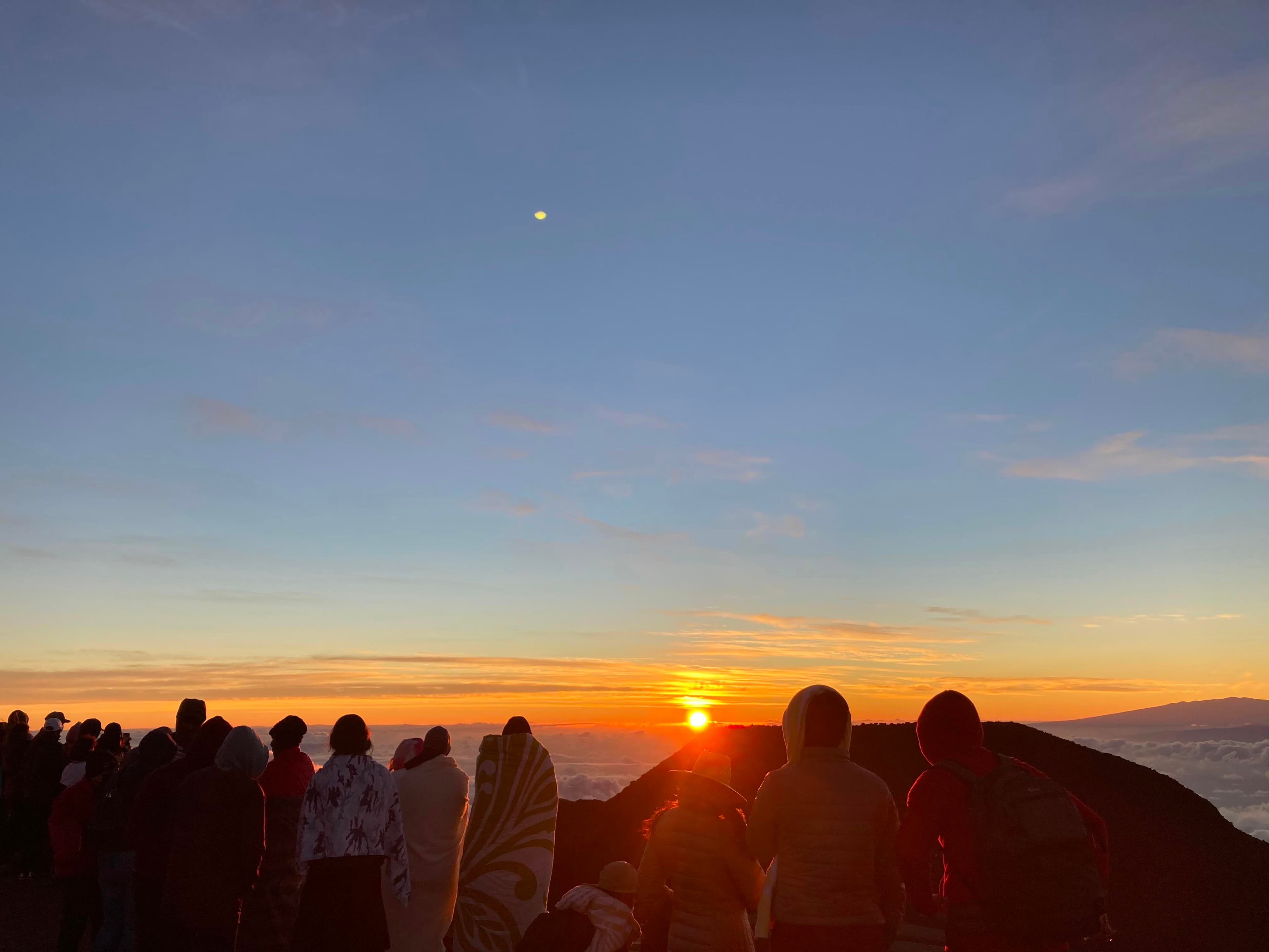 orange sun breaks horizon in distance people stand in blankets in foreground watching